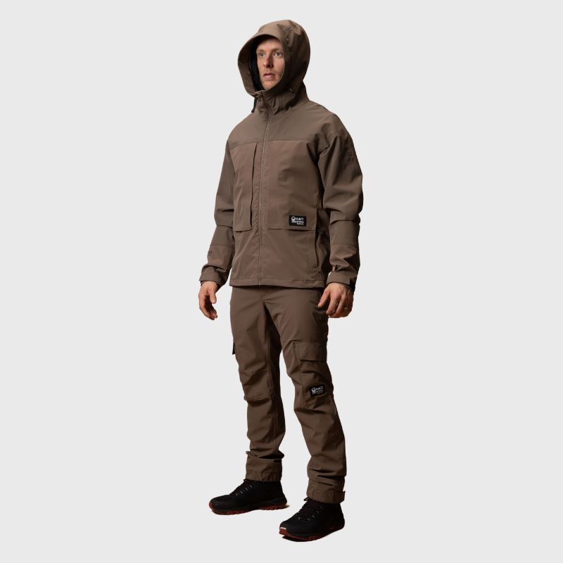 En person i brun Halti Hiker II DX Skaljacka Herr och matchande cargobyxor står mot en enfärgad ljusgrå bakgrund.