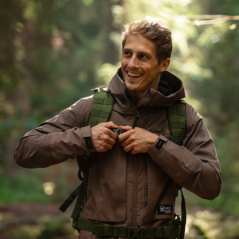 En person i en Halti Hiker II DX Skaljacka Herr och grön ryggsäck ler och justerar remmarna i en solbelyst skog.