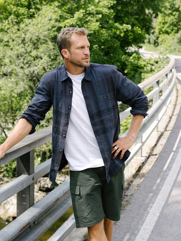 En man i en Halti Hiker Skjorta Herr och gröna shorts står på en bro, redo för friluftsaktiviteter bland träden.