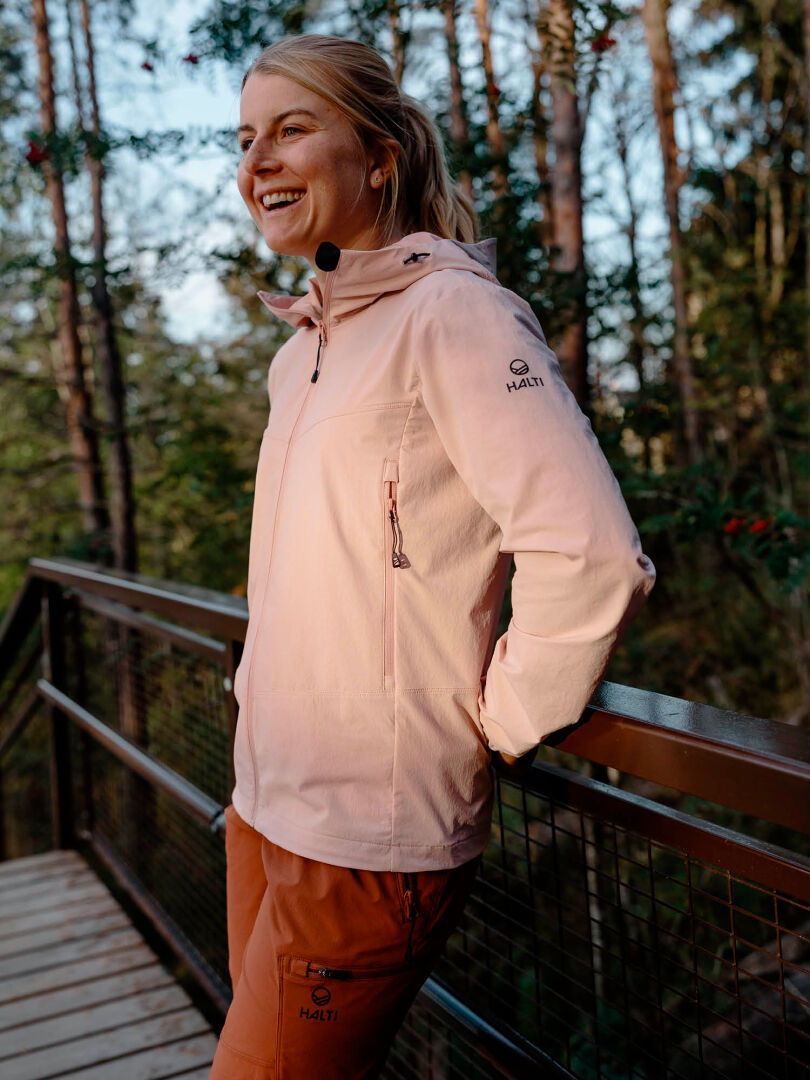 En person i lätt jacka och orange Halti Pallas Evo X-stretch Byxor Dam ler på en träbro i skogen.