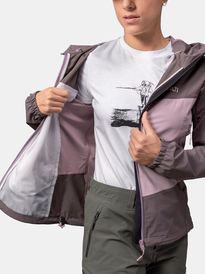 En person öppnar en Halti Pallas Evo X-stretch Jacka Dam och visar en vit grafisk t-shirt och gröna byxor mot en enfärgad bakgrund.
