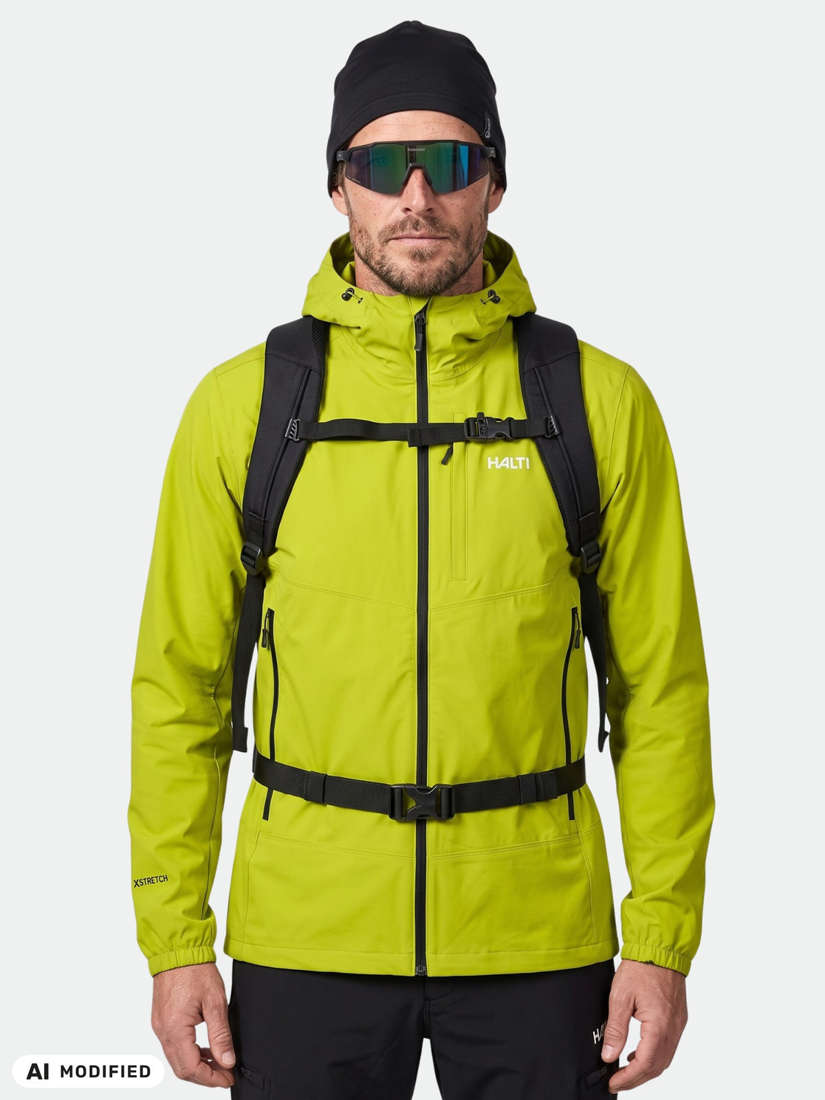 En man i en Halti Pallas Evo X-stretch-jacka för herrar, svarta byxor, mössa, solglasögon och ryggsäck – redo för en vandring.