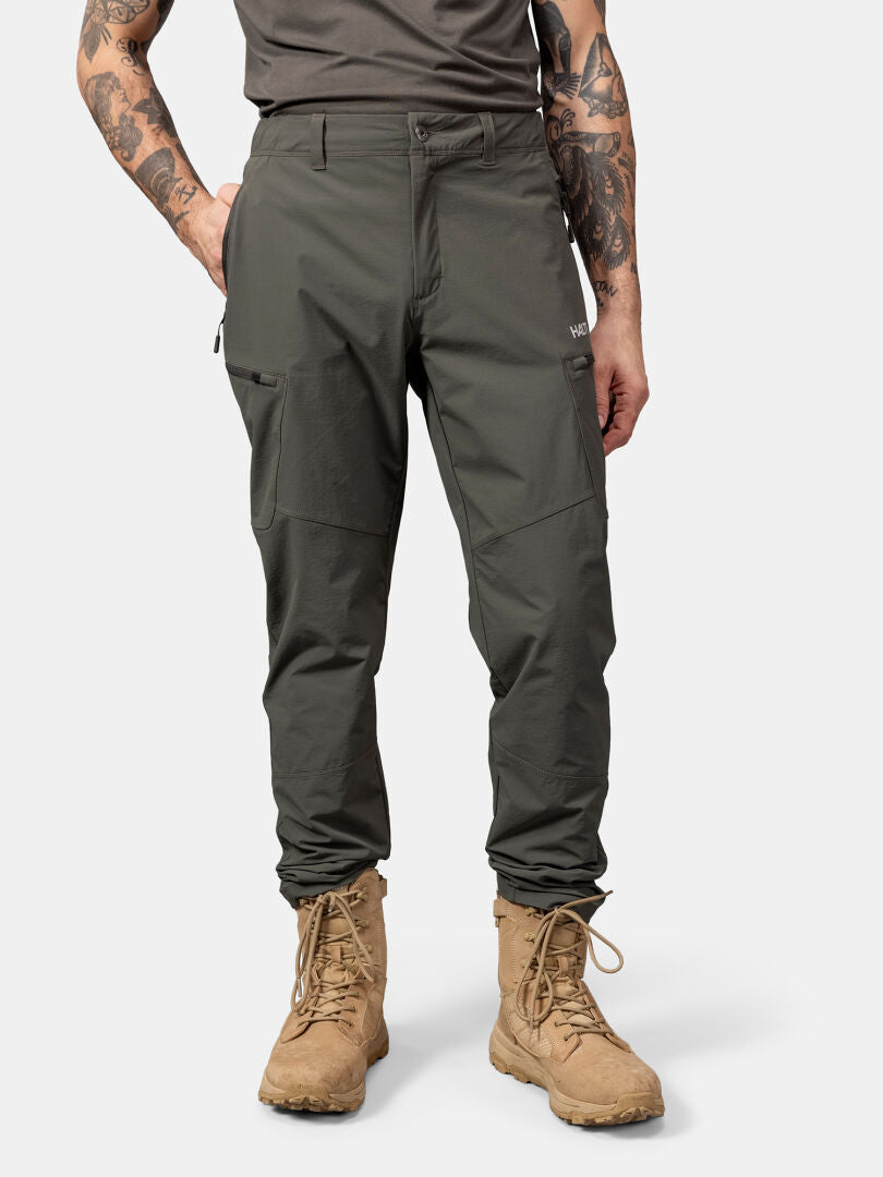 En person i Halti Pallas Evo X-stretch Byxor Herr och bruna snörkängor, med tatuerade armar, står mot en vit bakgrund.