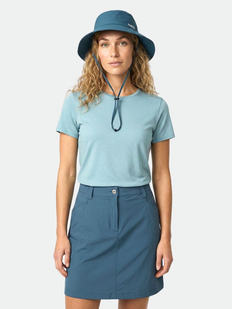 Kvinna i blågrön t-shirt, Halti Pallas Cool Skjol Dam-kjol och solhatt med hakrem, mot en enfärgad bakgrund.