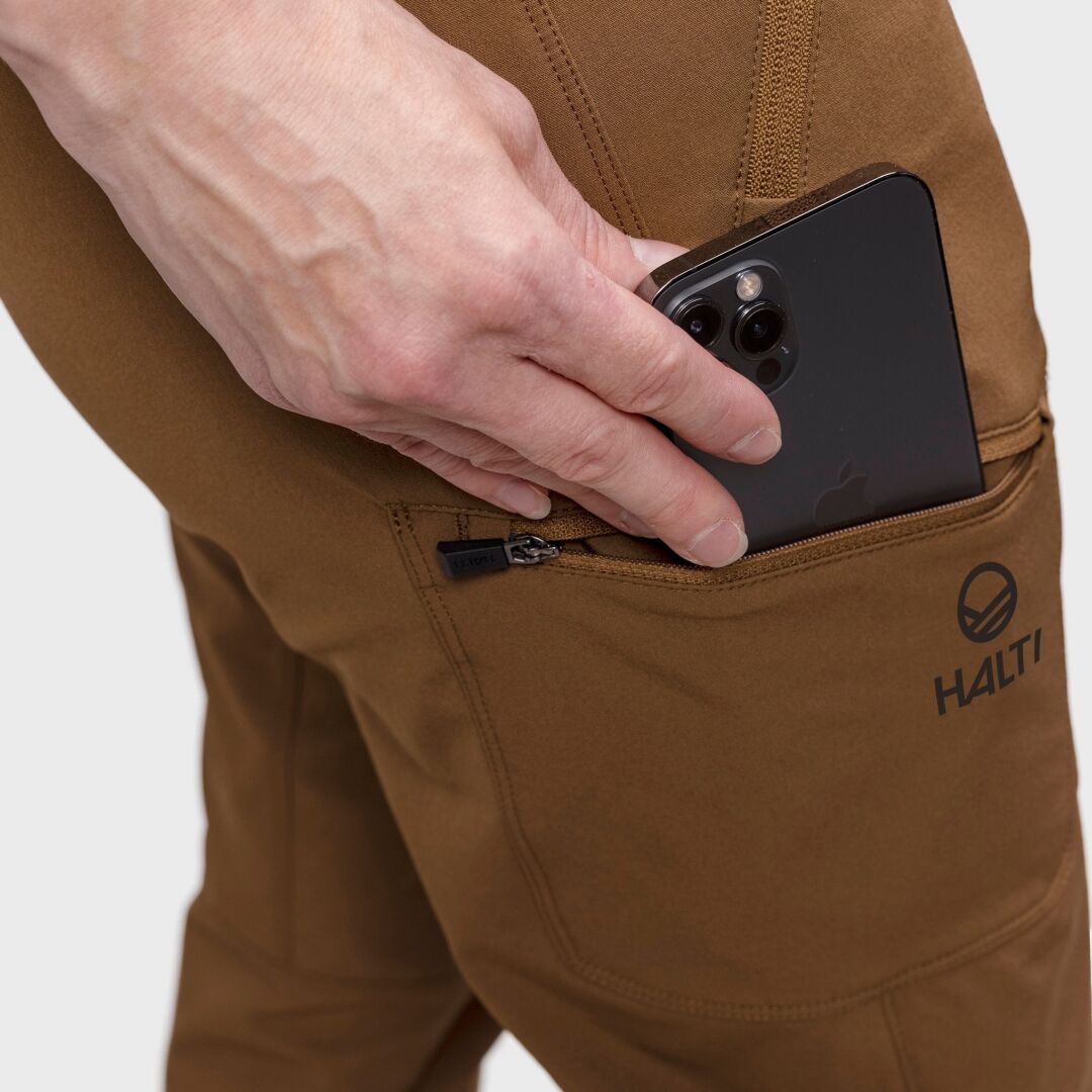 Någon stoppar en svart smartphone i den dragkedjeförsedda fickan på en brun Halti Pallas Evo Brushed X-stretch Byxor Dam.