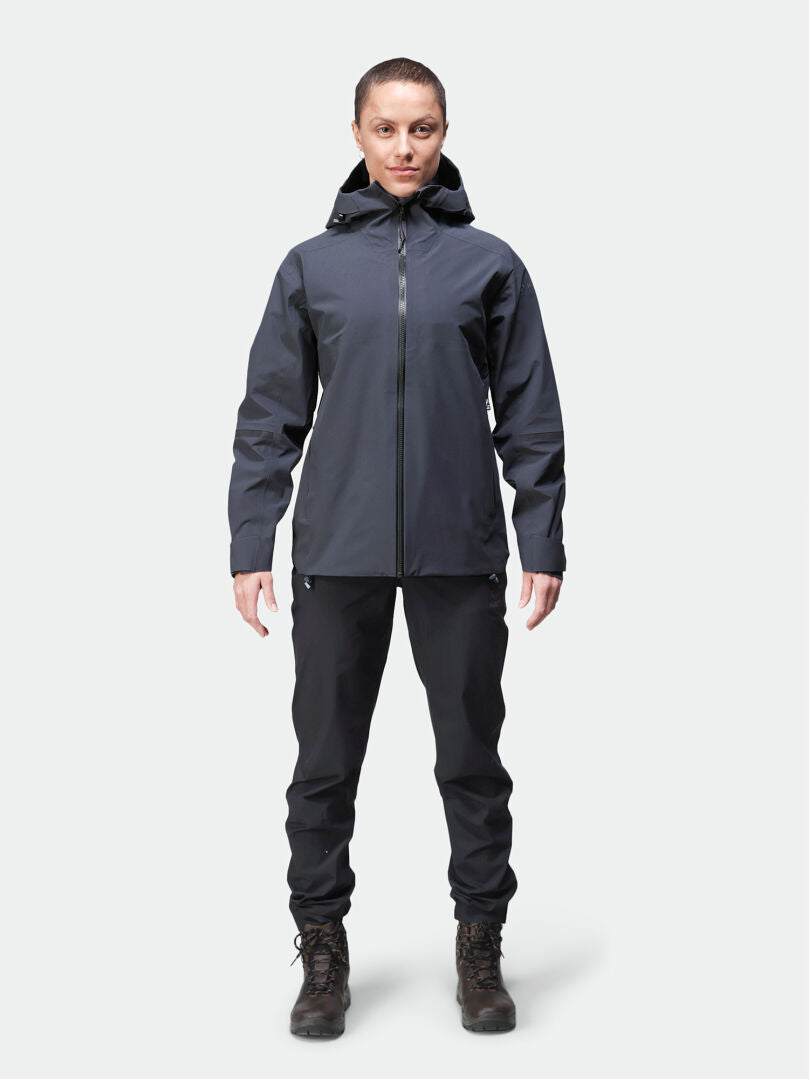 Person vänd framåt i en Halti Kajana 3L DX Skaljacka Dam, svarta byxor och vandringskängor mot en enfärgad bakgrund.