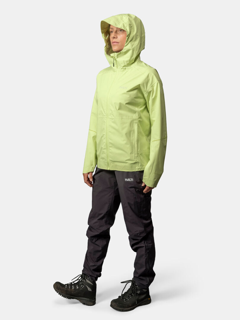 En person bär en ljusgrön Halti Pisarat 2,5L DX Skaljacka Dam, svarta byxor och vandringskängor mot en enfärgad bakgrund.