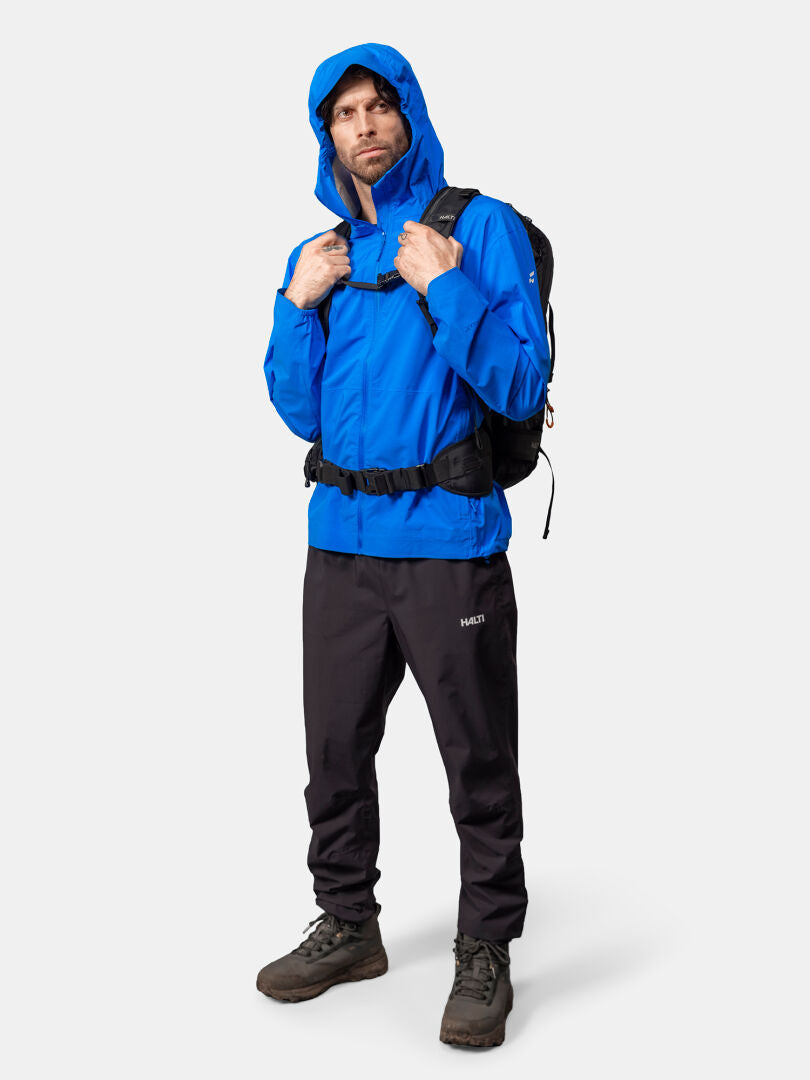 En man klädd i Halti Pisarat 2,5L DX Skaljacka Herr, svarta byxor och stövlar står med en svart ryggsäck.