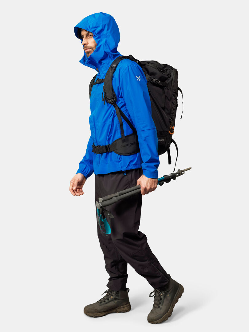 En person i en Halti Pisarat 2,5L DX Skaljacka Herr, svarta byxor, stövlar, med vandringsstavar och en svart ryggsäck.