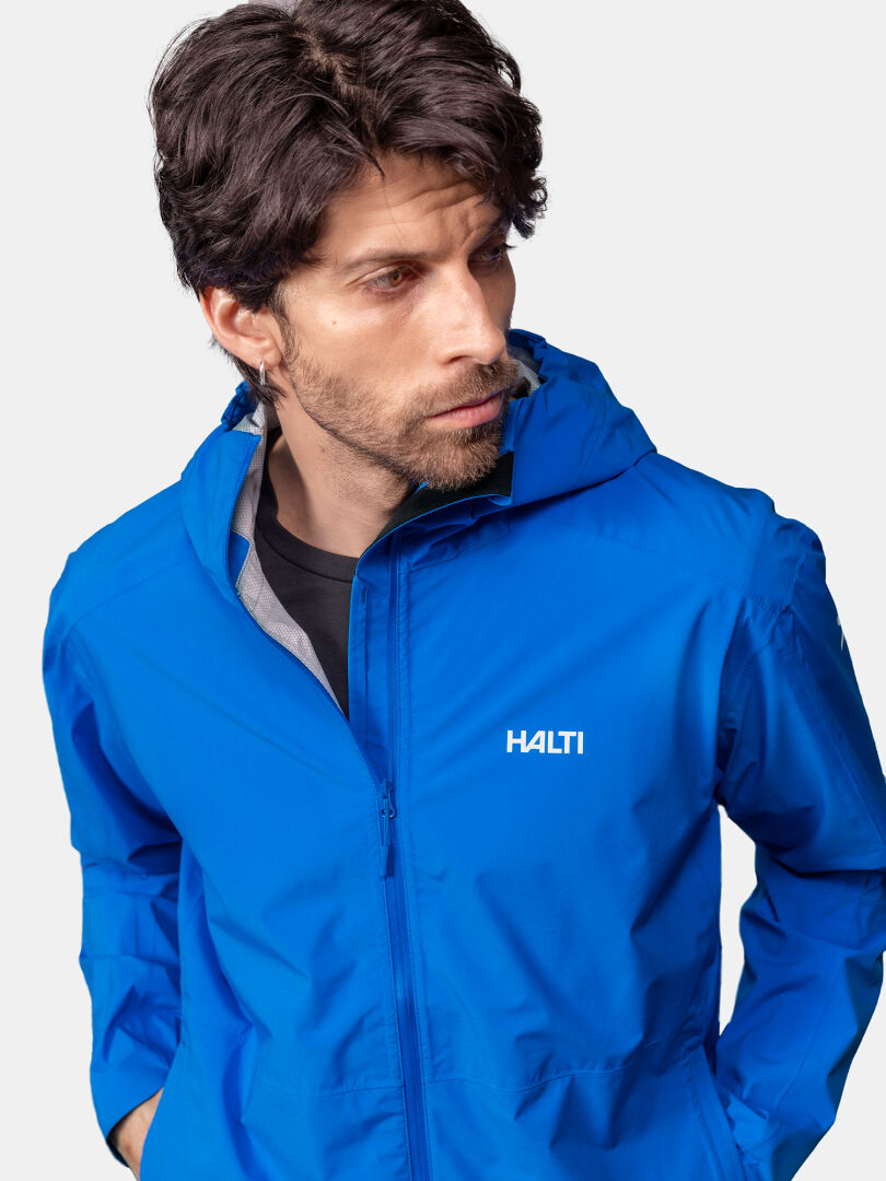 En man i en blå Halti Pisarat 2,5L DX Skaljacka Herr med huva tittar åt vänster mot en enfärgad ljus bakgrund.