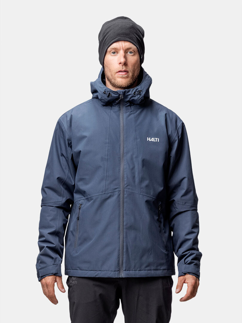 En man i mörkblå Halti Forter DX Warm Skaljacka Herr med huva och svart mössa står mot en enfärgad bakgrund.