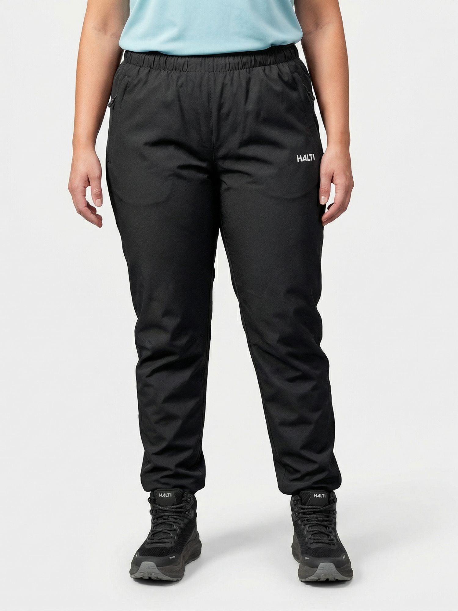 En person står vänd framåt i Halti Forter DX Skalbyxor Dam Plus och skor mot en enfärgad bakgrund.