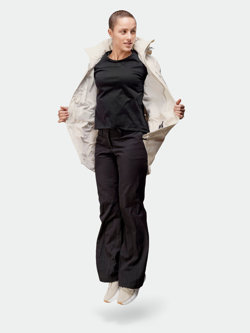 En person i svart skjorta, byxor och vita sneakers hoppar i en öppen Halti Kallas DX Parka Dam mot en enfärgad bakgrund.