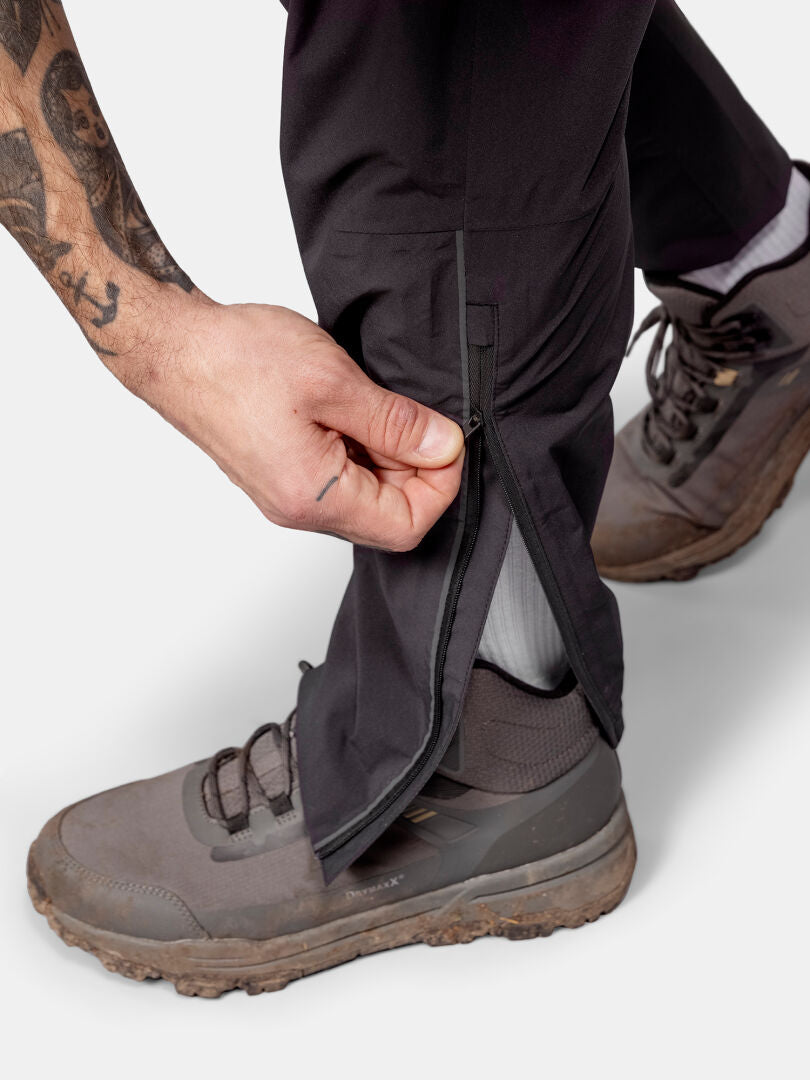 En person iförd grå vandringsskor drar upp dragkedjan på underbenet på Halti Pisarat 2,5L DX Skalbyxor Herr.