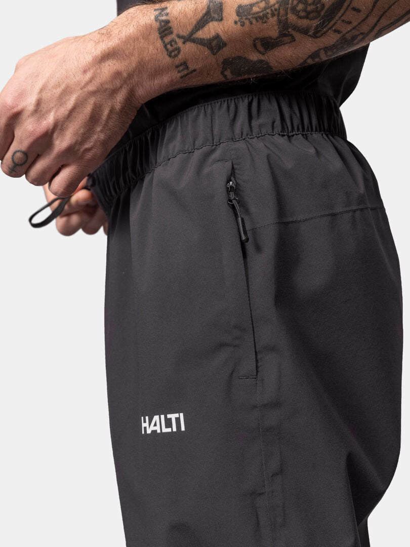 Närbild på en person som justerar Halti Pisarat 2,5L DX Skalbyxor Herr, visar tatuerad arm och ficka med dragkedja.