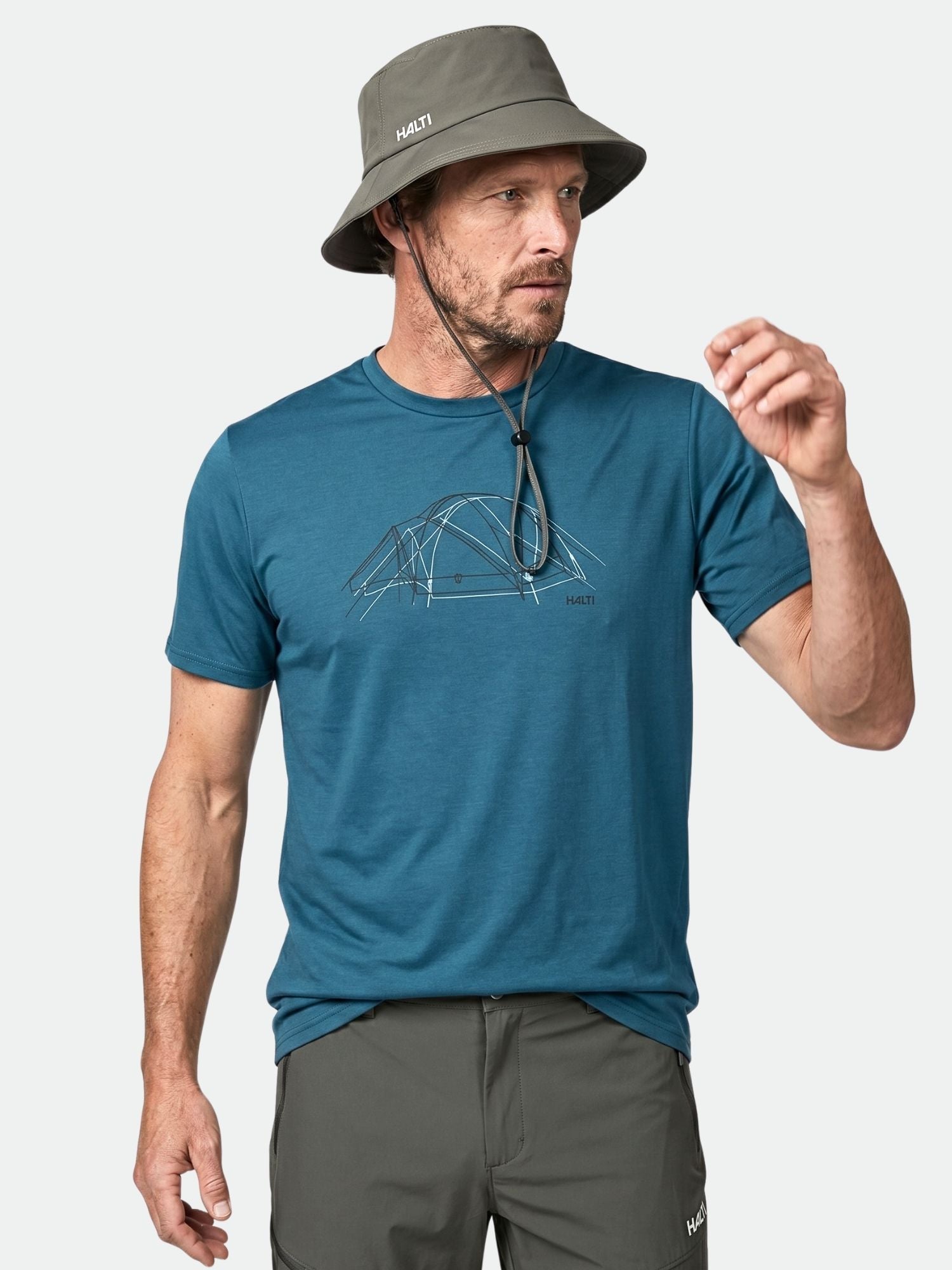 En man i Halti Kalteva T-Skjorta Herr med tältmotiv, grå hatt och grå byxor står mot en enfärgad bakgrund.