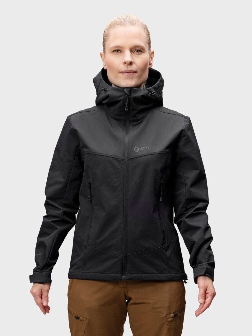 En person i svart Halti Pallas Evo Brushed X-stretch Jacka Dam och bruna byxor står mot en ljusgrå bakgrund.