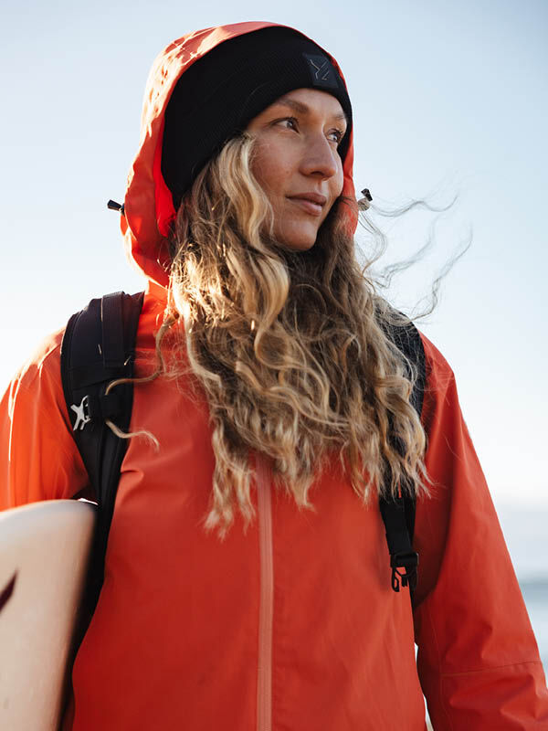Person med långt blont hår i Halti Staala DX Skaljacka Dam och svart hatt håller i en surfbräda utomhus.