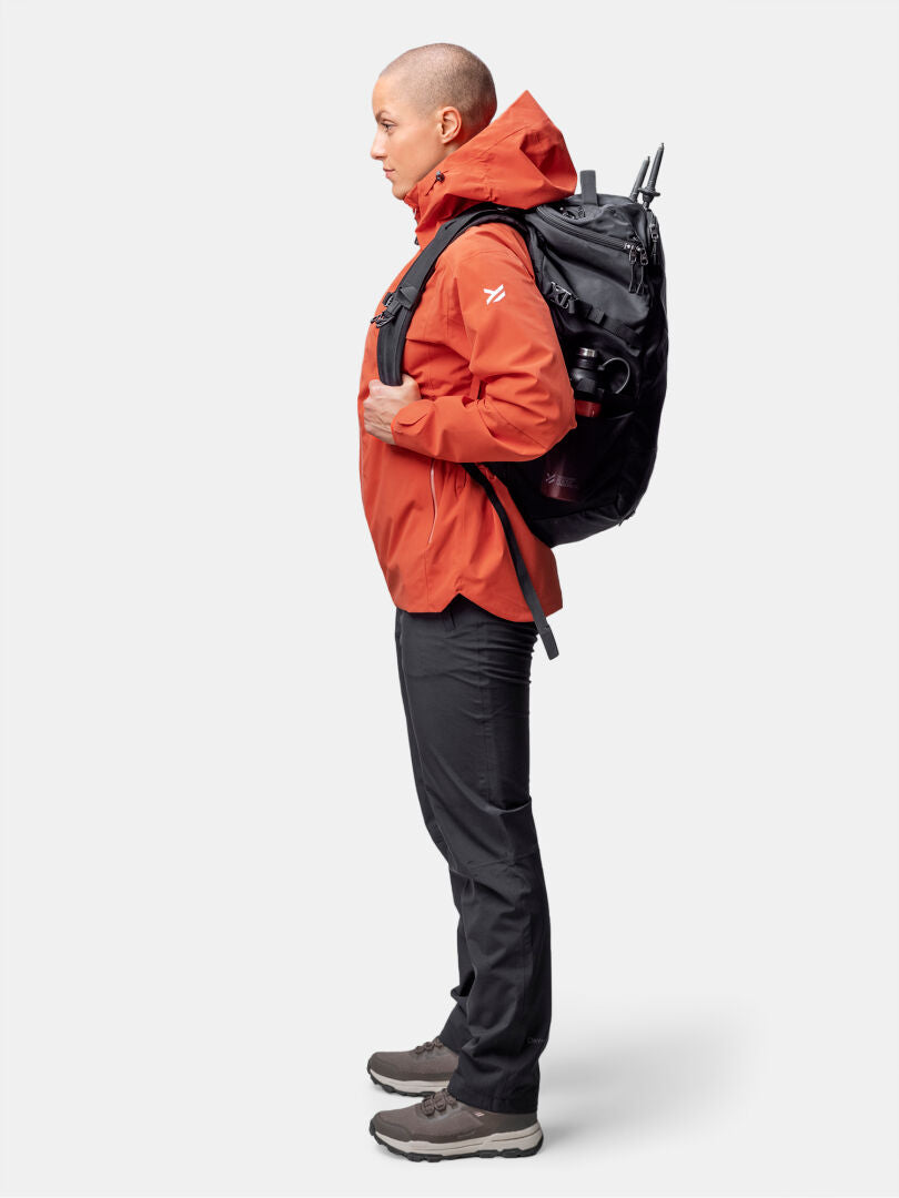 Personen i profil bär en Halti Staala DX Skaljacka Dam (orange), svarta byxor, bruna skor och en stor svart ryggsäck.