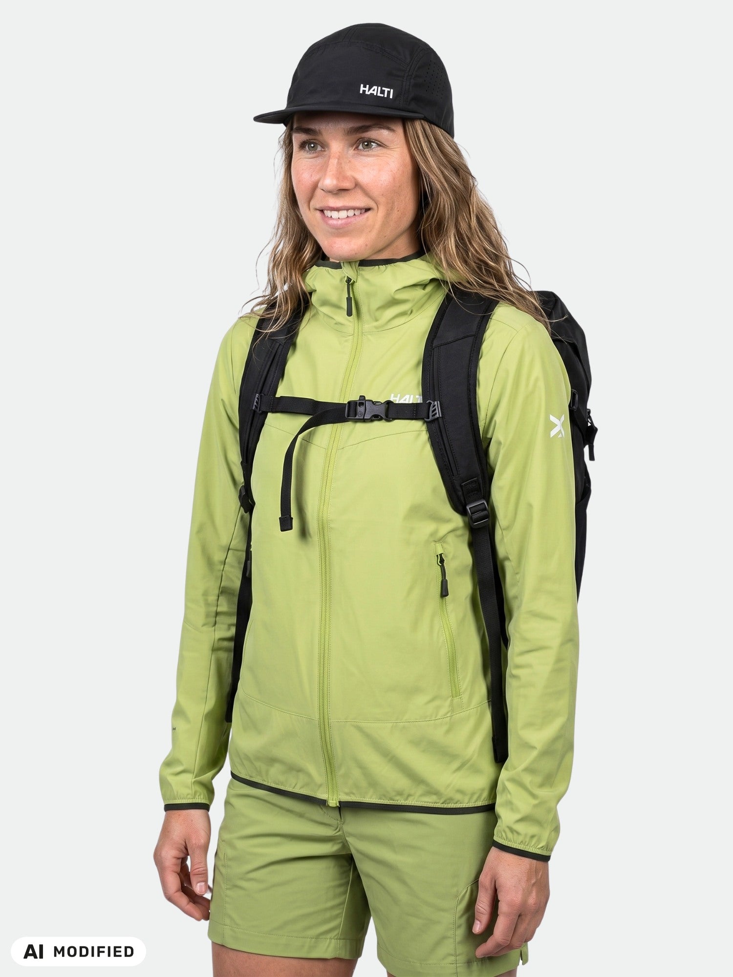 En person bär en Halti Pallas Cool X-stretch-jacka för dam (ljusgrön), shorts, en svart ryggsäck och en keps, framför en enfärgad bakgrund.