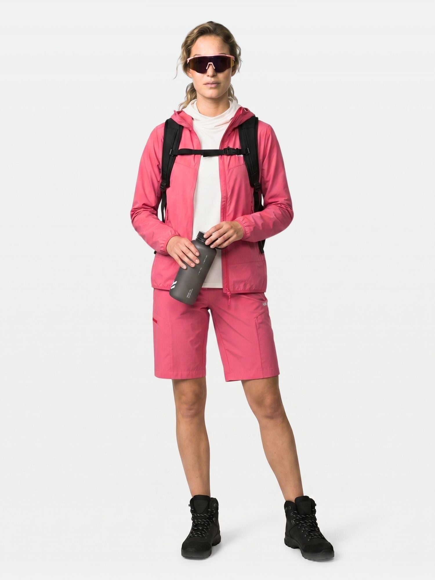 En person i rosa Halti Pallas Cool X-stretch jacka och svarta stövlar står och håller i en tumbler, bär ryggsäck och solglasögon.