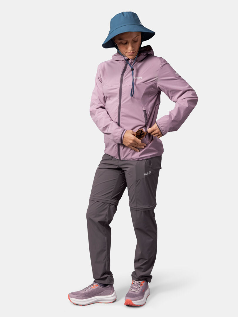 Person i blå hatt, grå byxor och Halti Pallas Cool X-stretch Jacka Dam håller i solglasögon mot en vit bakgrund.
