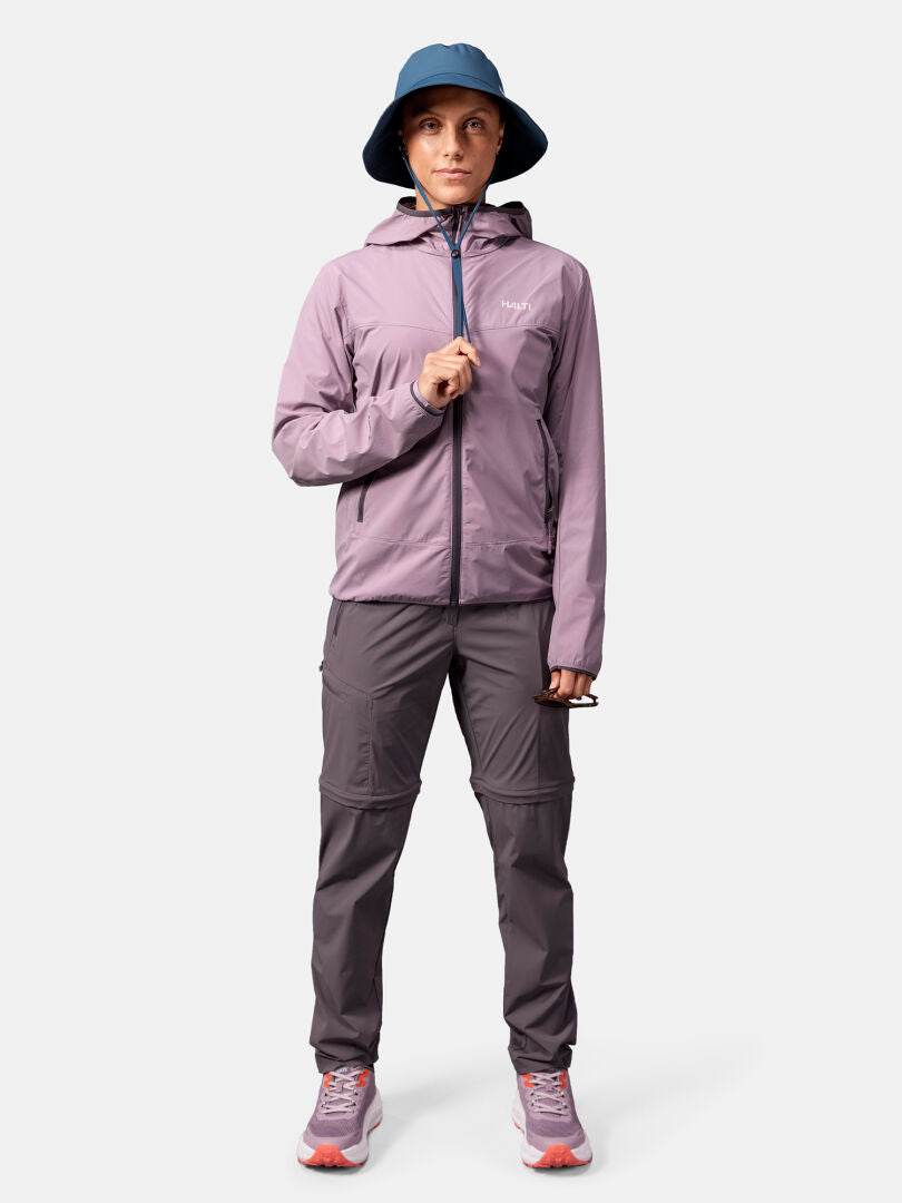 Person i lila Halti Pallas Cool X-stretch Jacka Dam, grå byxor, blå hatt och rosa sneakers står mot en enfärgad bakgrund.