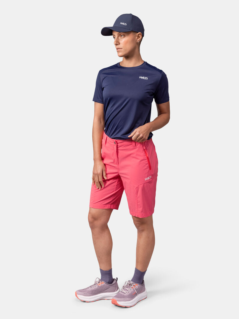 Kvinna i Halti Pallas Cool Shorts Dam, sneakers, marinblå t-shirt och keps - perfekt för friluftsliv mot en ljus bakgrund.