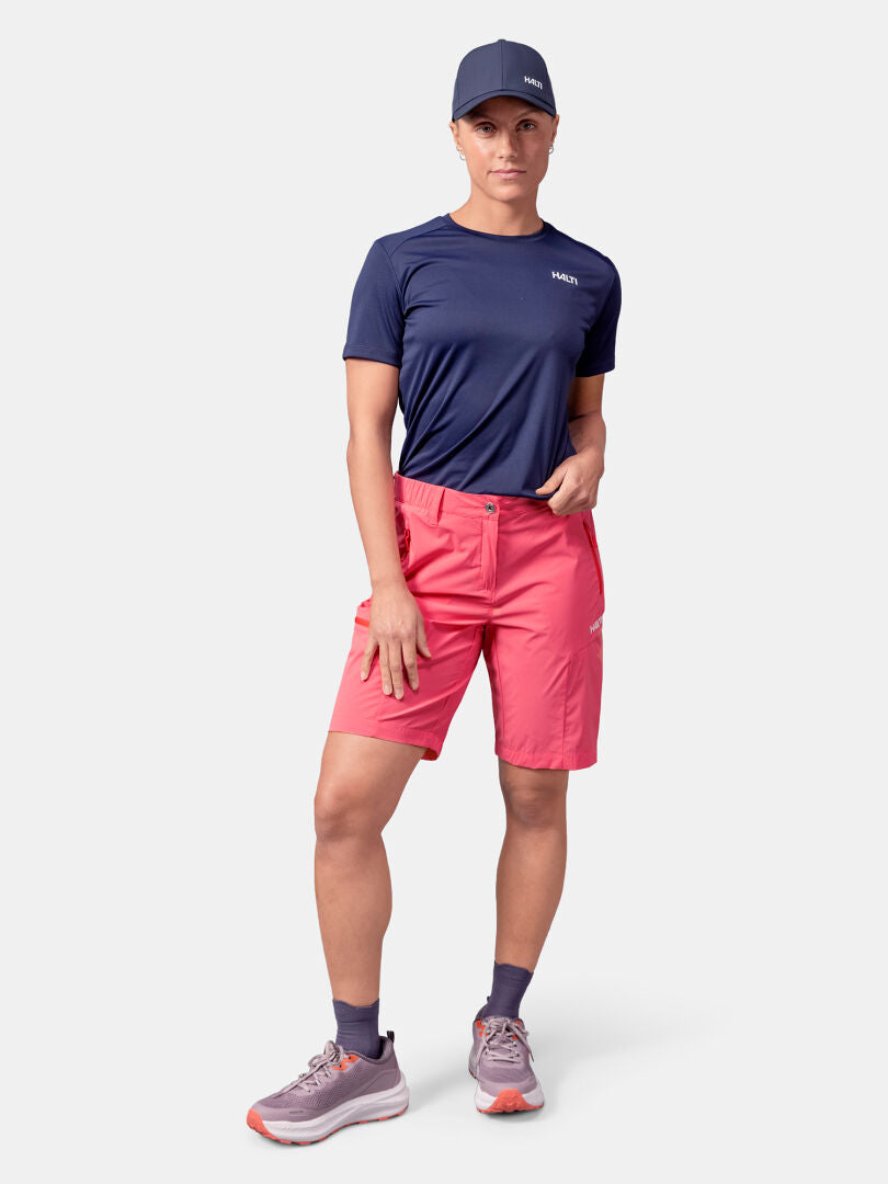 Klädd i Halti Pallas Cool Shorts Dam, marinblå t-shirt och keps - redo för friluftsliv mot en enfärgad bakgrund.