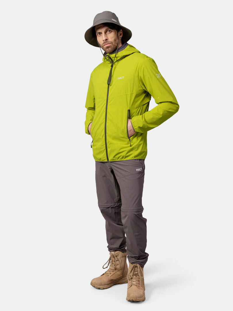En person klädd i Halti Pallas Cool X-stretch Jacka Herr, grå byxor, bruna stövlar och en grå bredbrättad hatt står med händerna i fickorna.