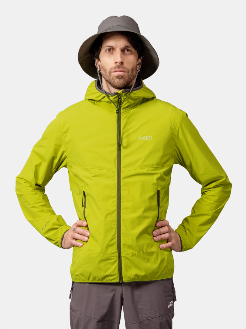 En man bär en Halti Pallas Cool X-stretch Jacka Herr i limegrönt och en grå hatt, stående mot en vit bakgrund.