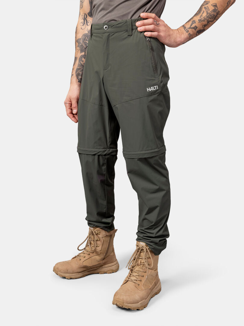 Man i Halti Pallas Cool Stretch Zip-off Byxor Herr, solbruna stövlar och kortärmad skjorta mot enfärgad bakgrund.