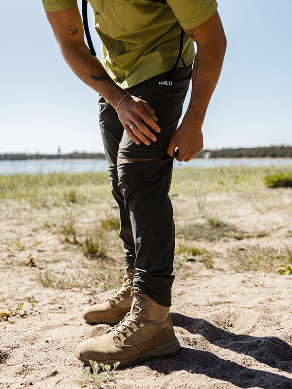 Person utomhus på sand, iklädd Halti Pallas Cool Stretch Zip-off Byxor Herr med UPF 90+, en hand på knät.