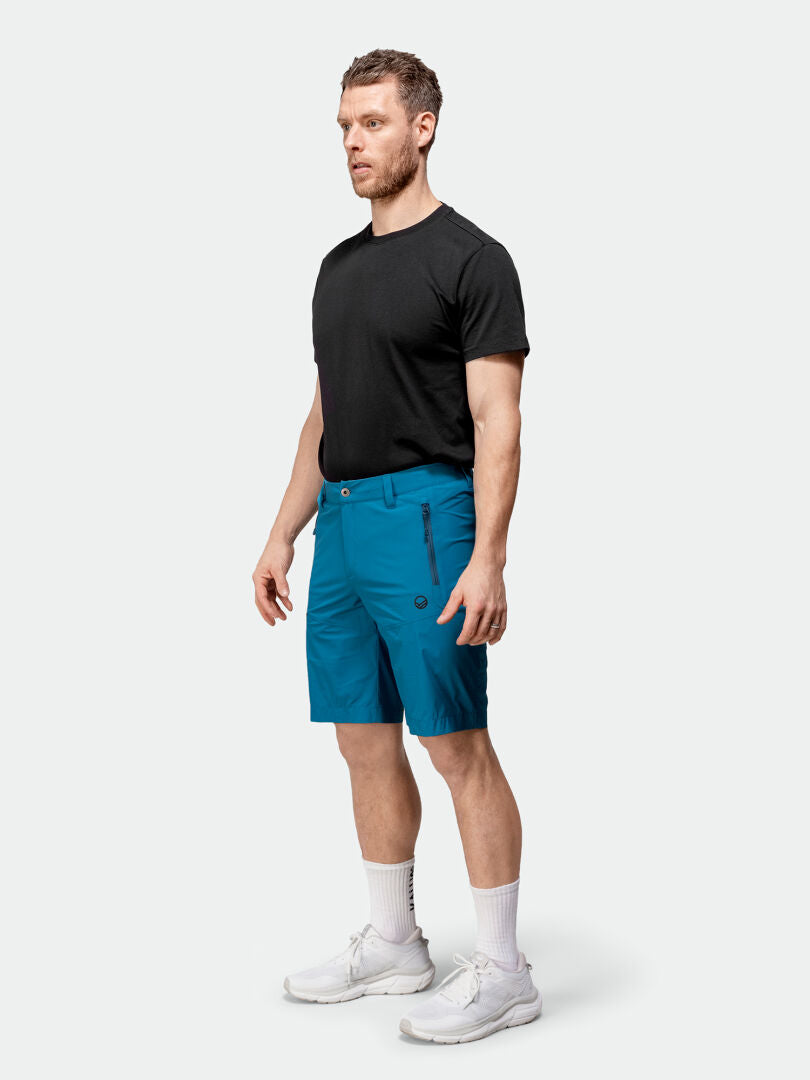 En man vetter mot vänster i en svart t-shirt och blå Halti Pallas Cool X-stretch Shorts Herr, med vita strumpor och sneakers.