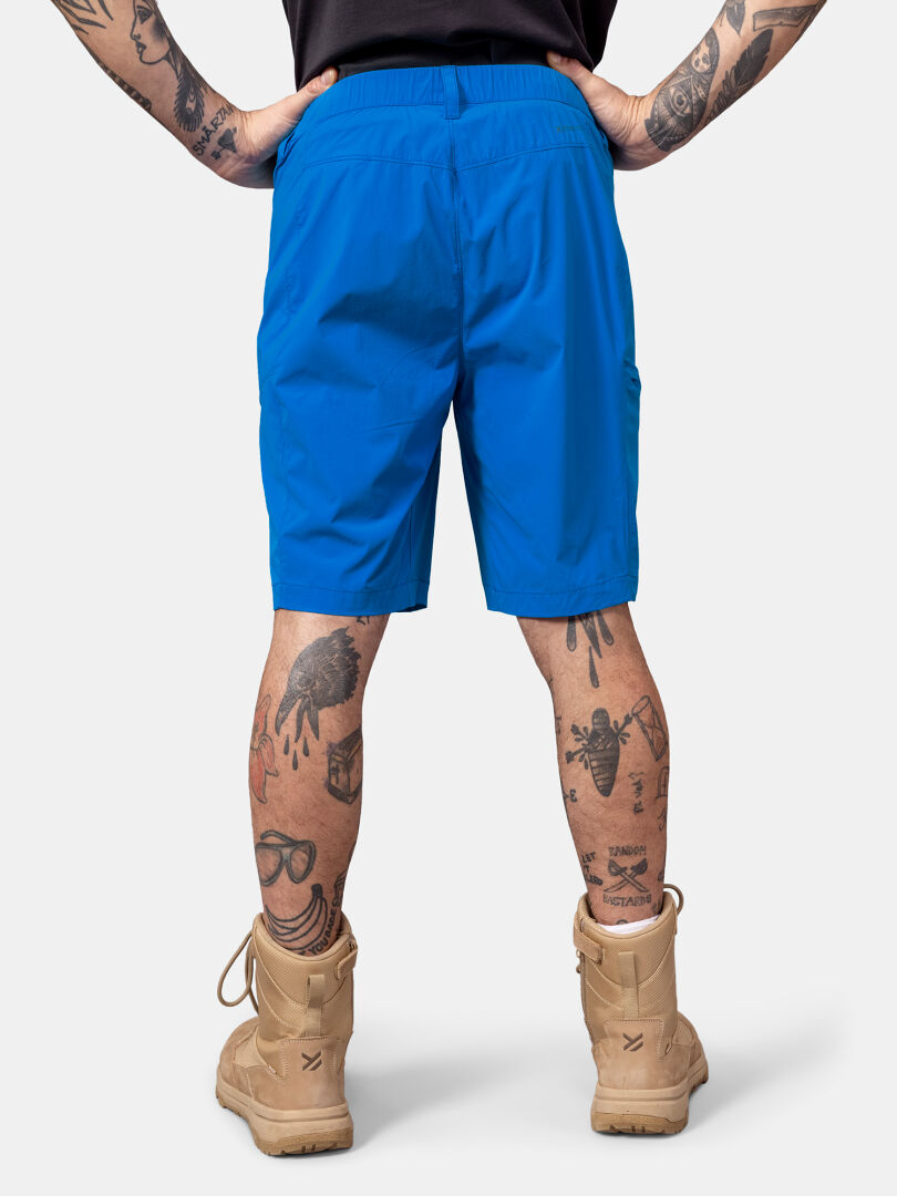 Person klädd i Halti Pallas Cool X-stretch Shorts, solbruna stövlar, svart skjorta; tatueringar på ben och armar synliga, fotograferad bakifrån.