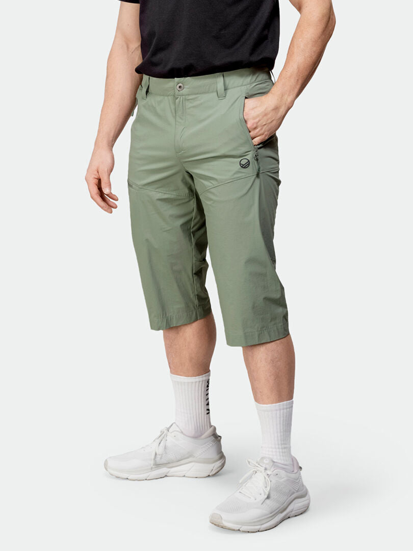 En person i Halti Pallas Cool X-stretch Capribyxor Herr, en svart t-shirt, vita strumpor och sneakers står mot en enfärgad bakgrund.