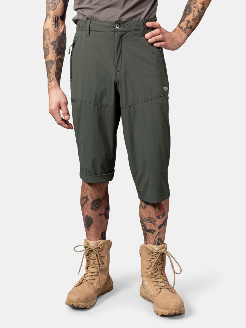En man i Halti Pallas Cool X-stretch Capribyxor, grå skjorta och solbruna kängor står mot en enfärgad bakgrund.