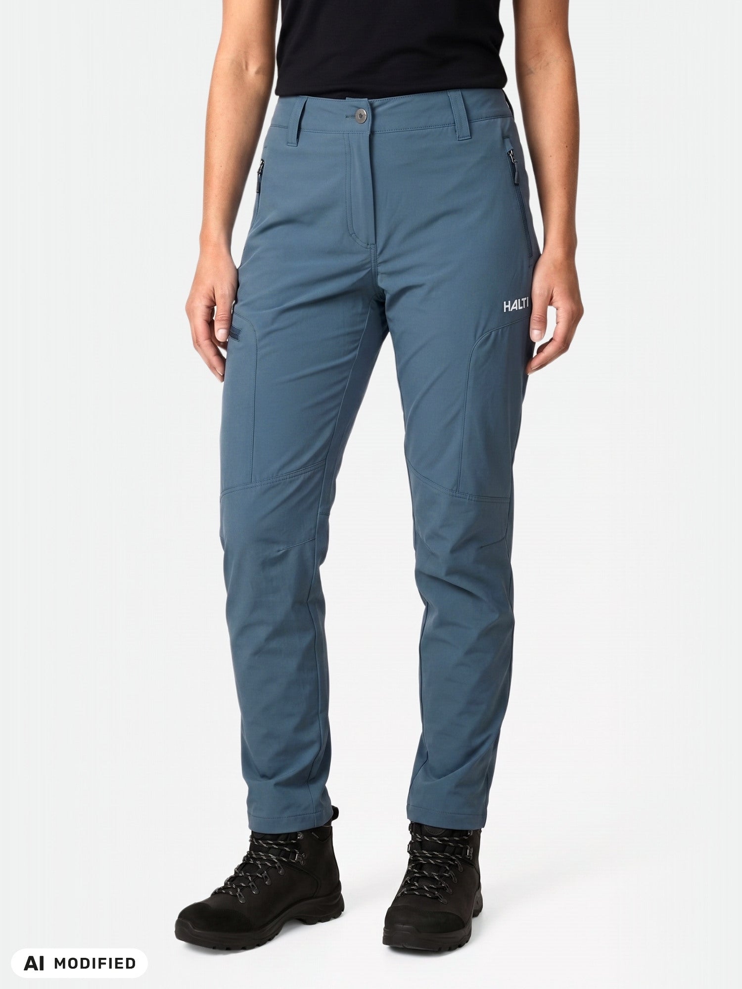 En kvinna i vandringsbyxor av modellen Halti Pallas Cool X-stretch, svarta stövlar och en skjorta, stående mot en vit bakgrund.