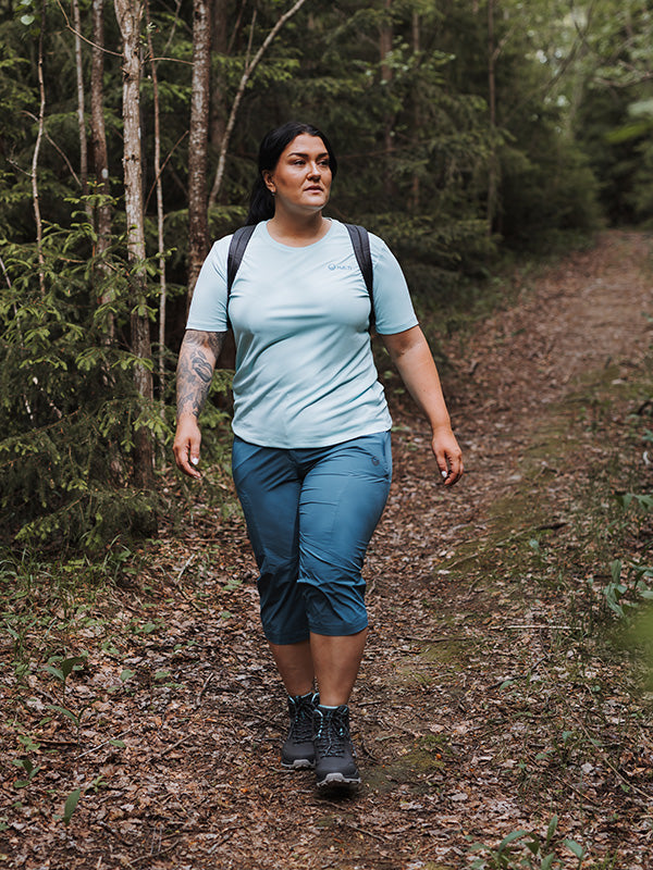 Iklädd Halti Pallas Cool X-stretch Capribyxor går en vandrare med ryggsäck på en skogsstig.