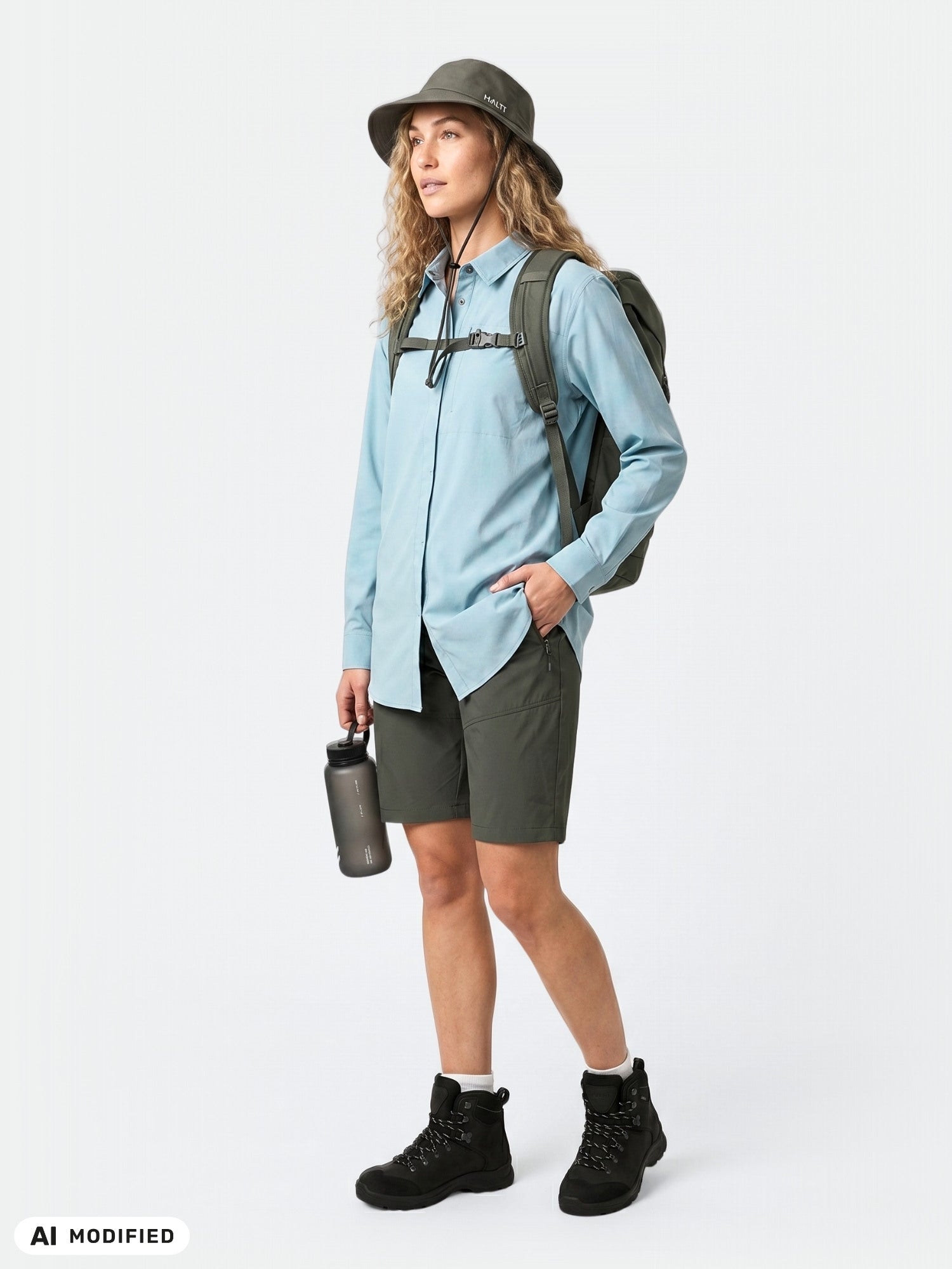 En person klädd i en Halti Samoy långärmad funktionsskjorta för damer, gröna shorts, stövlar och mössa står med en ryggsäck och en flaska.