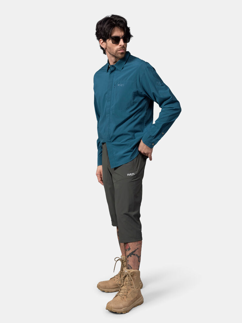 En man i blågrön Halti Samoy Långärmad Funktionsskjorta, olivfärgade shorts och bruna stövlar står vänd lite åt vänster.