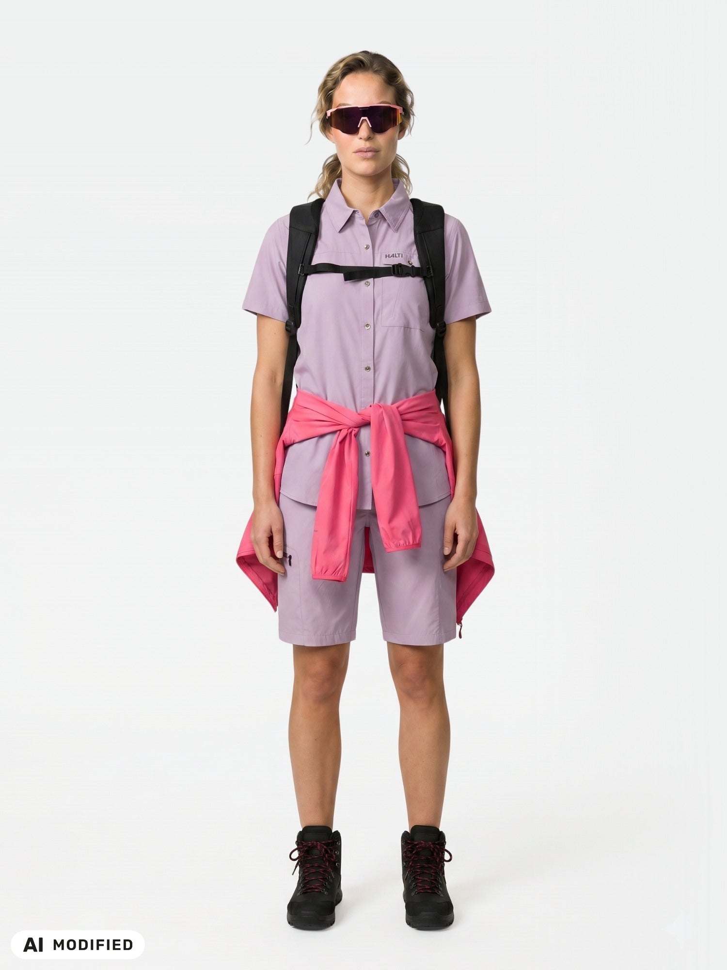 Kvinna i Halti Pallas Cool Shorts Dam, ljuslila, med en rosa jacka knuten runt midjan, svarta stövlar och solglasögon.