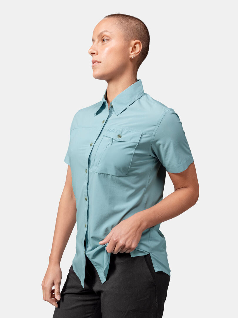 Person med kort hår i ljusblå Halti Samoy Funktionsskjorta Dam och svarta byxor, vänd åt vänster på enfärgad bakgrund.
