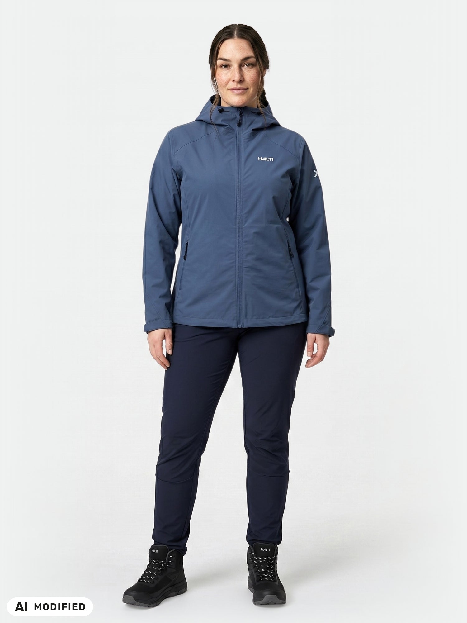En person klädd i en blå Halti Finholma X-stretch Jacka Dam Plus, mörka byxor och svarta stövlar står framför en vit bakgrund.