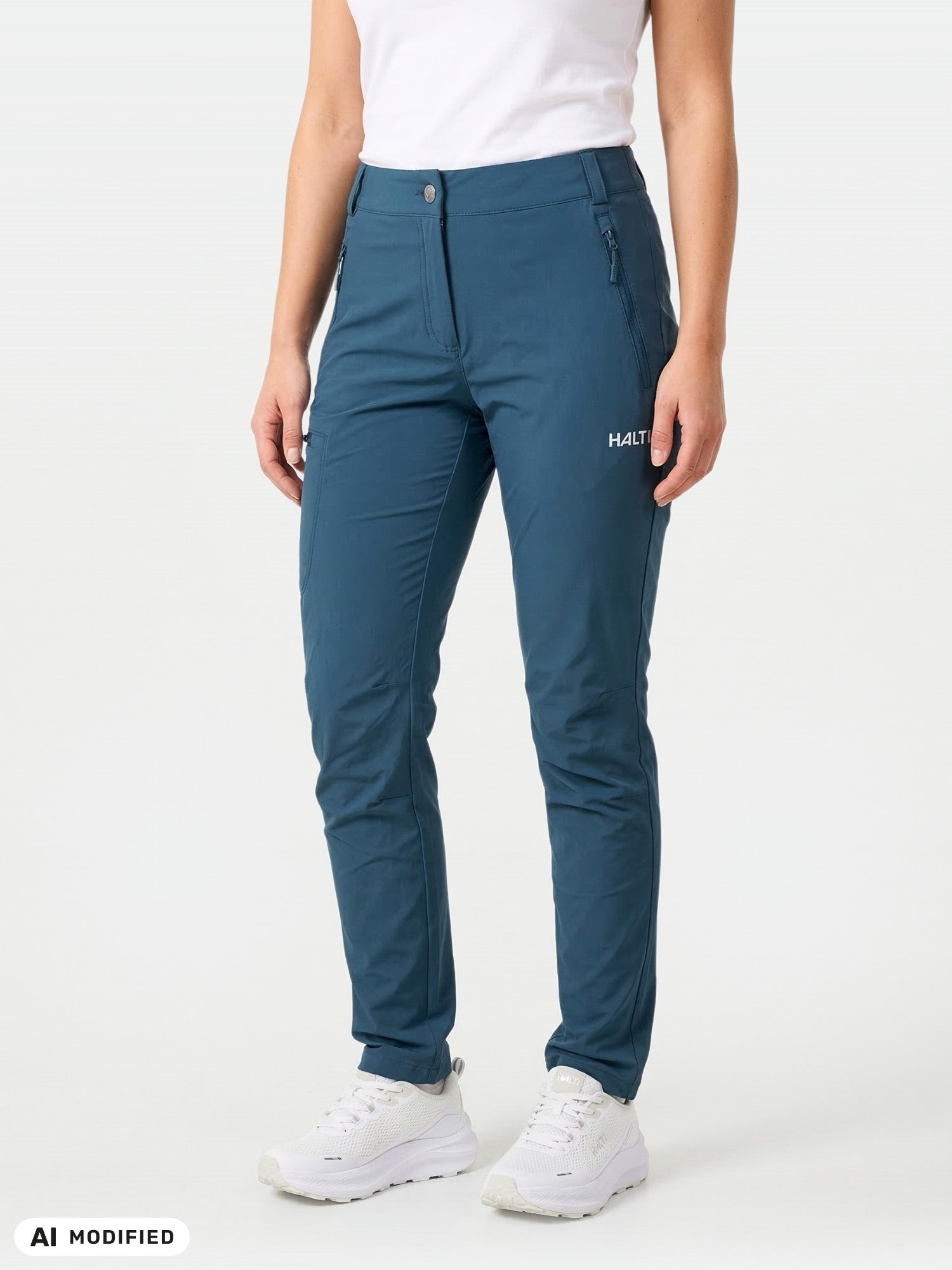 En kvinna i turkosa Halti Pallas X-stretch Outdoor-byxor, vita sneakers och en skjorta, som står vid en ljus vägg.