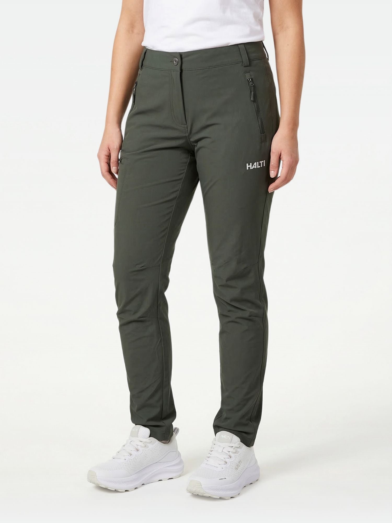 En person klädd i Halti Pallas X-stretch Outdoor Byxor Dam, vit skjorta och vita sneakers står mot en enfärgad bakgrund.