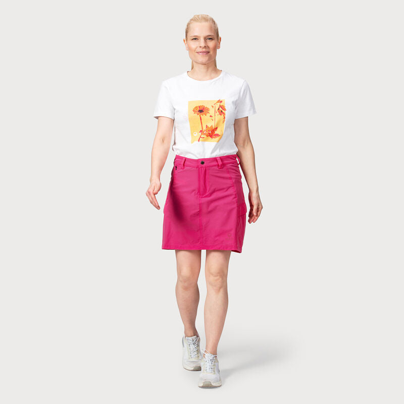 En kvinna går fram i en vit t-shirt med mönster, en Halti Reissu Dam Stretch Kjol-kort och sneakers mot en enfärgad bakgrund.