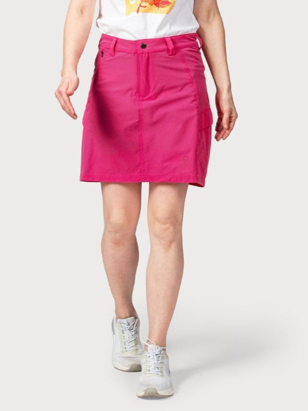 En person bär en ljusrosa Halti Reissu Dam Stretch Kjol, vit grafisk T-shirt och vita sneakers mot en enfärgad bakgrund.