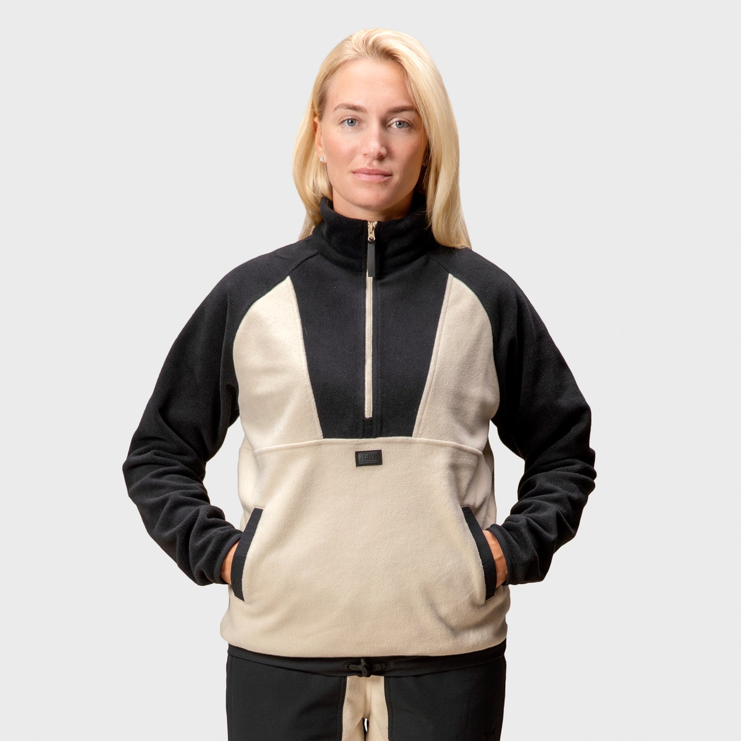 En kvinna visar Halti Skugge Unisex Fleece Anorak med händerna i framfickan, vilket framhäver dess mångsidiga design.