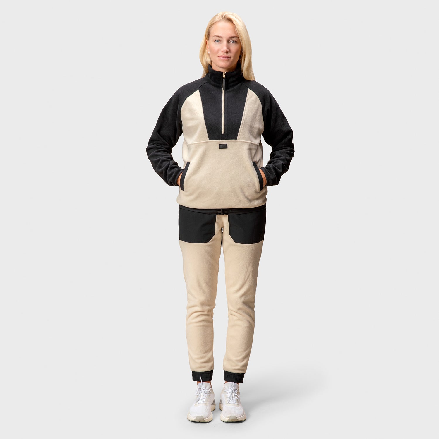 En kvinna står fram i en Halti Skugge Unisex fleece-anorak och byxor, och stylar en elegant athleisure-jacka med vita sneakers.